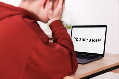 Photo of Cyber bullying. Upset teenage boy and laptop with text You Loser at desk indoors, closeup