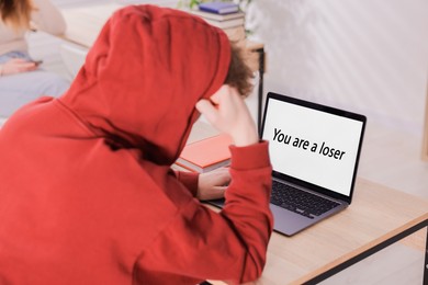 Photo of Cyber bullying. Upset teenage boy and laptop with text You Loser at desk indoors, closeup