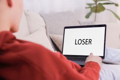 Photo of Cyber bullying. Upset teenage boy looking at laptop with text You Loser on sofa at home, closeup