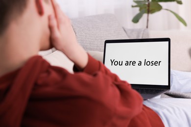 Photo of Cyber bullying. Upset teenage boy looking at laptop with text You Loser on sofa at home, closeup