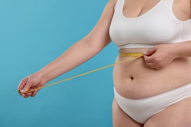 Photo of Weight loss. Plus size woman measuring waist with tape on light blue background, closeup