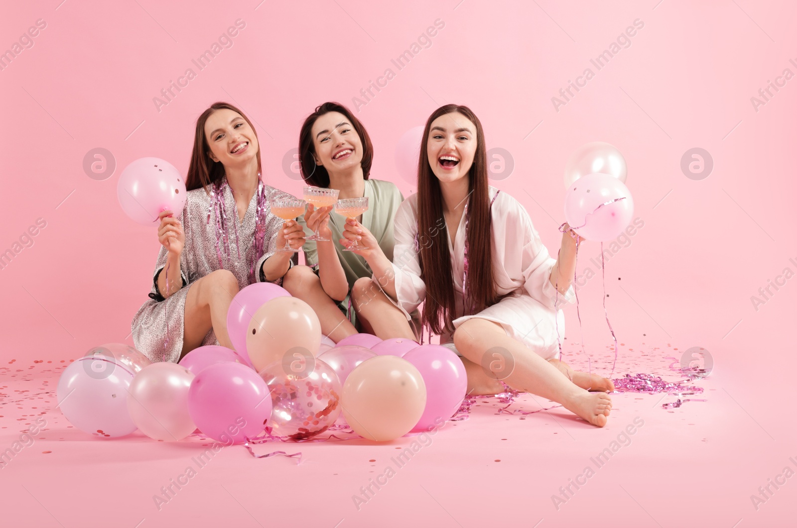 Pajama party. Happy friends with tinsel and balloons clinking glasses on pink background Photo of Pajama party. Happy friends with tinsel and balloons clinking glasses on pink background