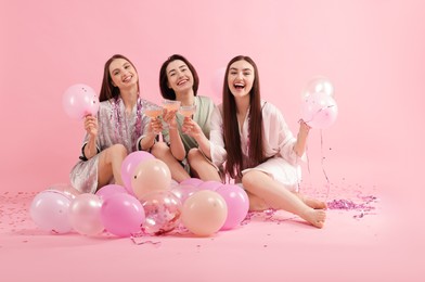 Photo of Pajama party. Happy friends with tinsel and balloons clinking glasses on pink background