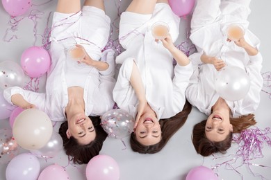 Photo of Pajama party. Happy friends with glasses of alcoholic drink, balloons and tinsel on grey background, top view