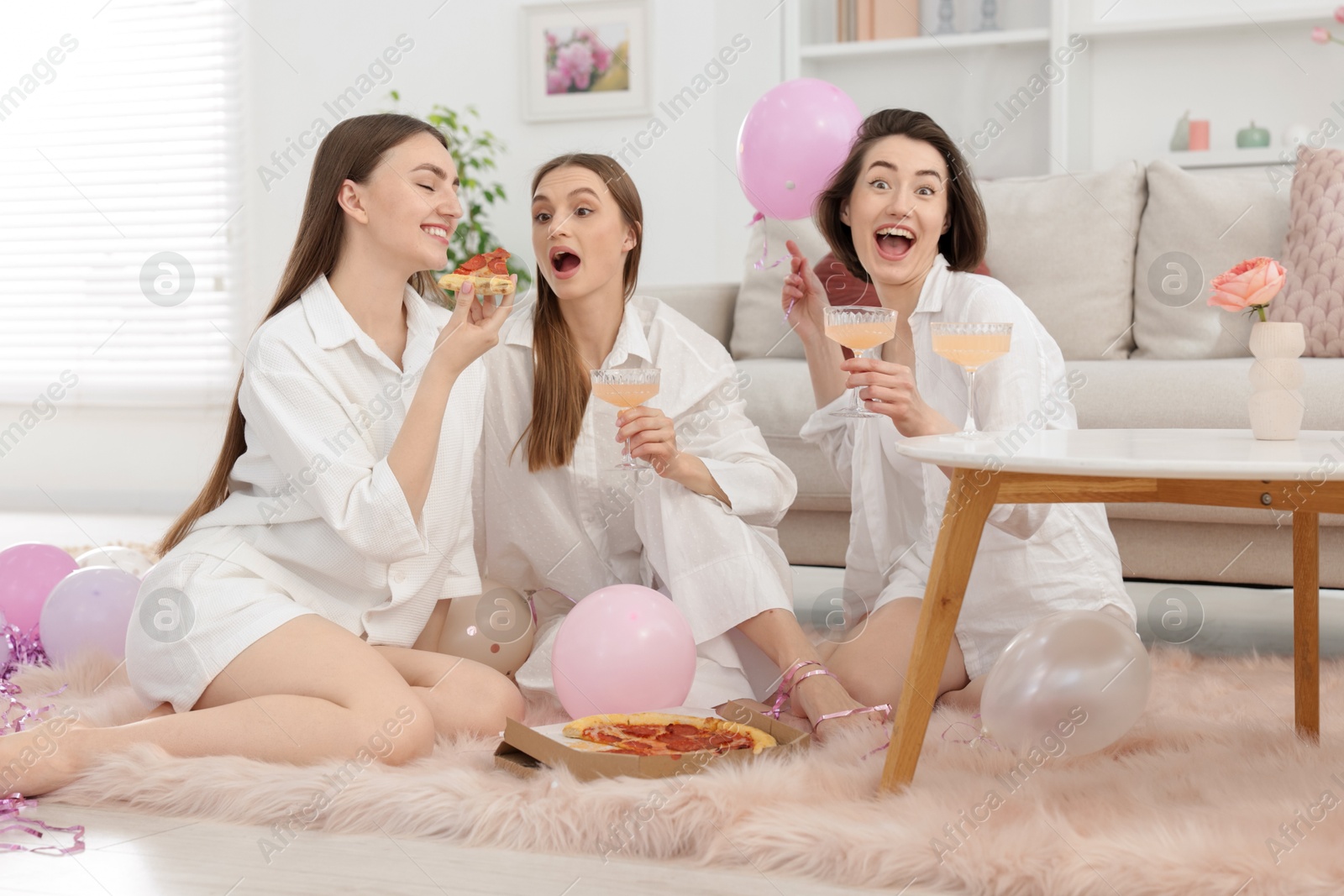 Happy friends in pajamas with glasses of alcoholic drink eating pizza on floor at home Photo of Happy friends in pajamas with glasses of alcoholic drink eating pizza on floor at home