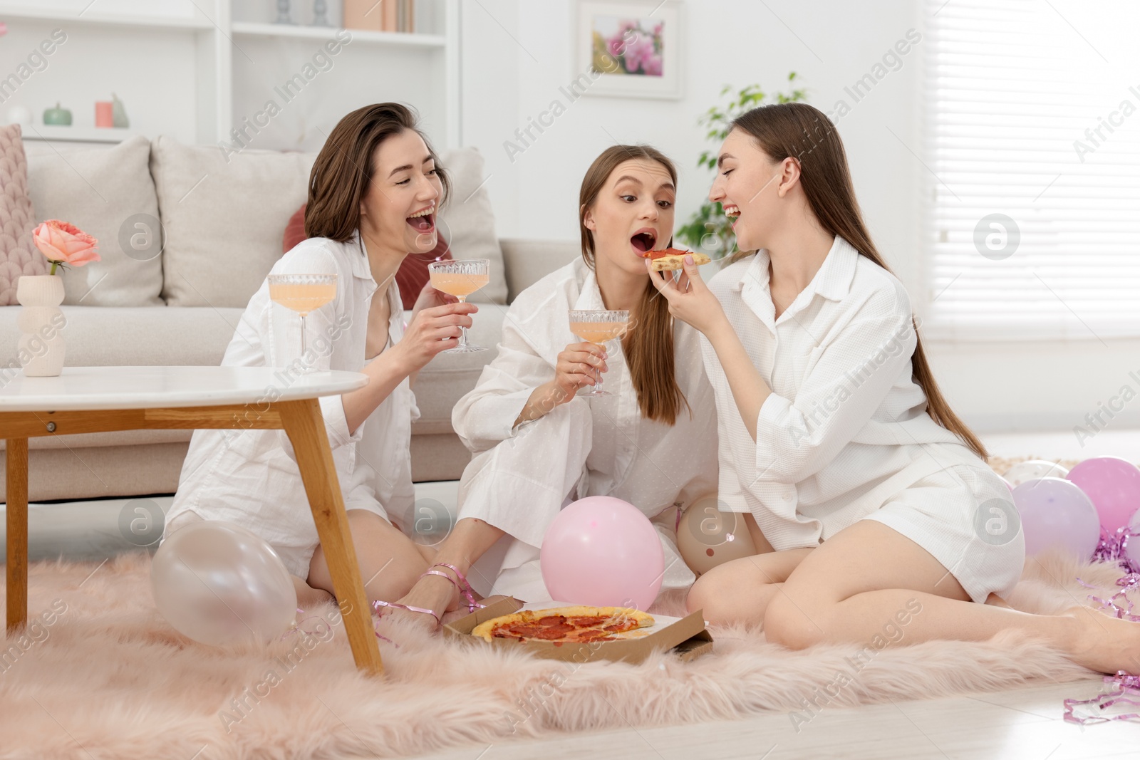 Happy friends in pajamas with glasses of alcoholic drink eating pizza on floor at home Photo of Happy friends in pajamas with glasses of alcoholic drink eating pizza on floor at home