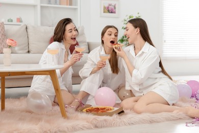Photo of Happy friends in pajamas with glasses of alcoholic drink eating pizza on floor at home
