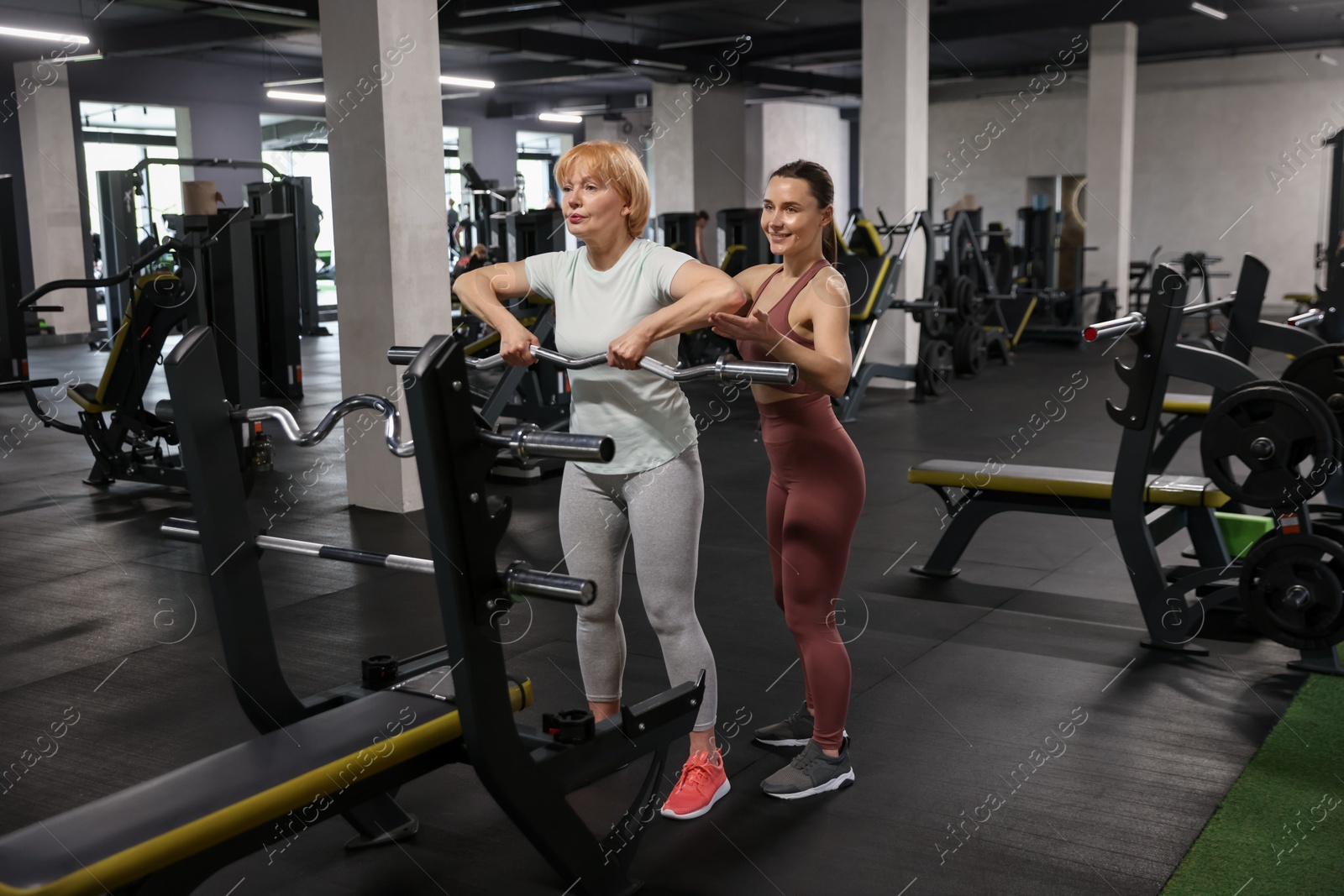 Senior woman exercising with EZ curl bar supervised by professional trainer in gym Photo of Senior woman exercising with EZ curl bar supervised by professional trainer in gym