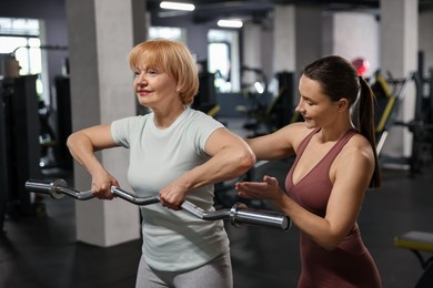 Photo of Senior woman exercising with EZ curl bar supervised by professional trainer in gym