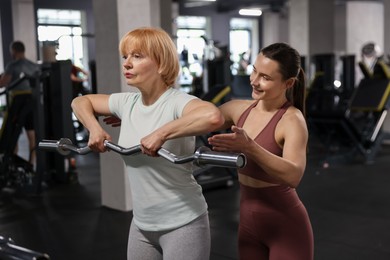 Photo of Senior woman exercising with EZ curl bar supervised by professional trainer in gym