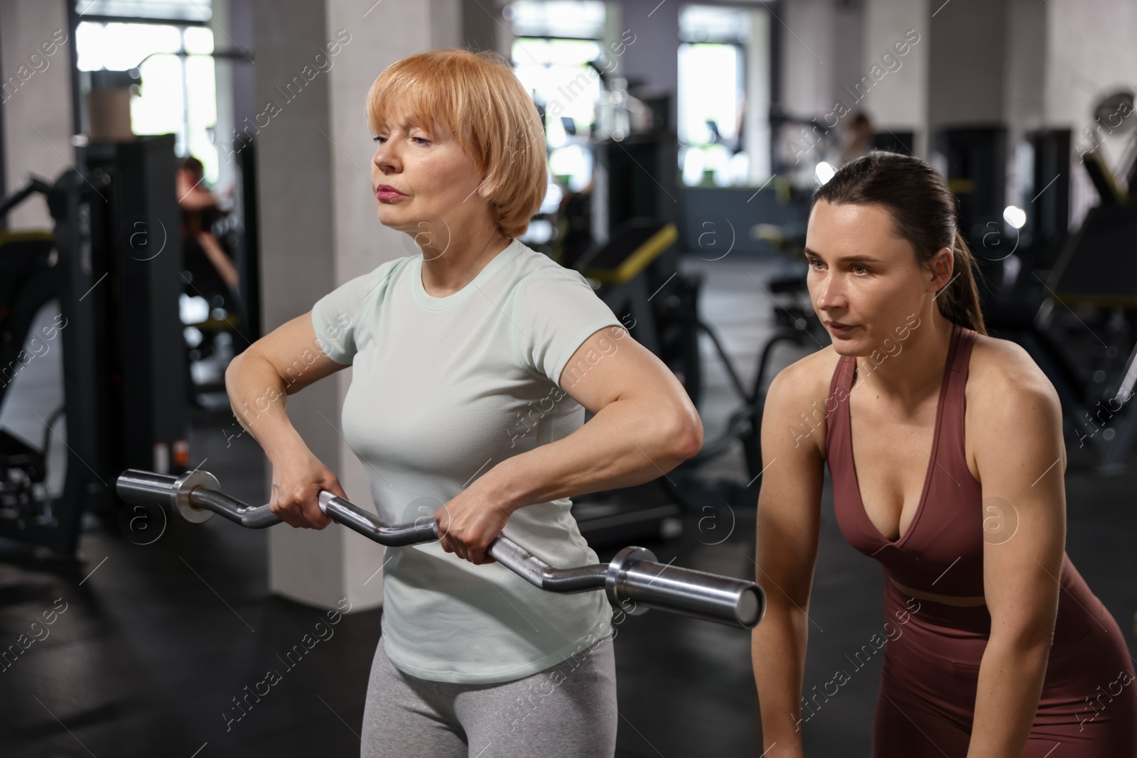Senior woman exercising with EZ curl bar supervised by professional trainer in gym Photo of Senior woman exercising with EZ curl bar supervised by professional trainer in gym