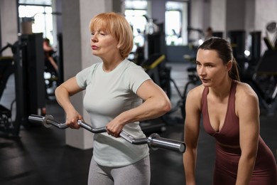 Photo of Senior woman exercising with EZ curl bar supervised by professional trainer in gym