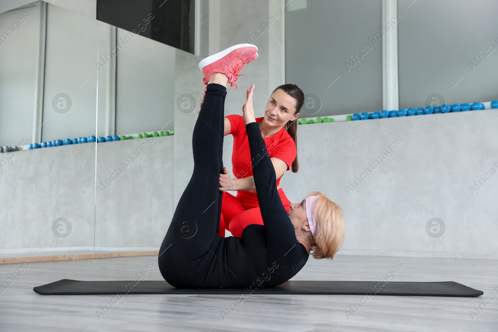 Senior woman exercising on yoga mat supervised by professional trainer in gym Photo of Senior woman exercising on yoga mat supervised by professional trainer in gym