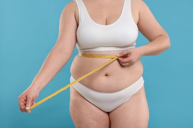 Photo of Weight loss. Plus size woman measuring waist with tape on light blue background, closeup