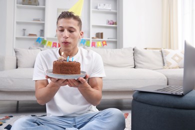 Photo of Birthday celebration. Man blowing candles on cake while having video call via laptop at home