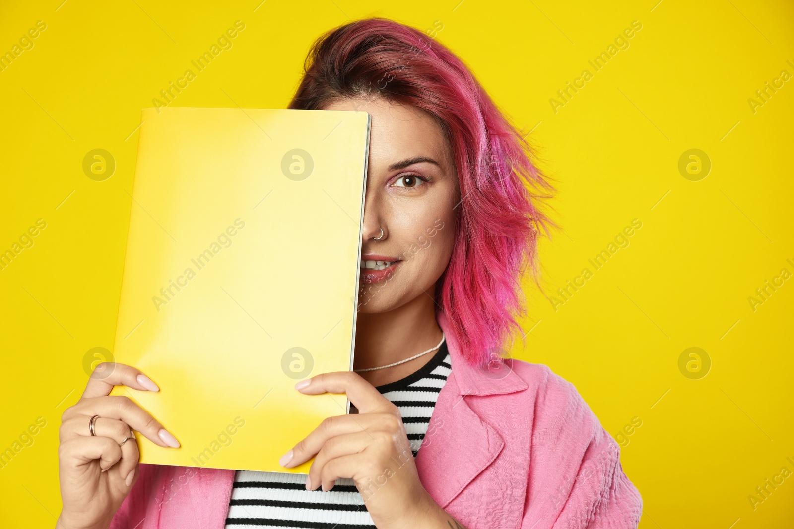 Beautiful woman with pink dyed hair covering eye with fashion magazine on yellow background Photo of Beautiful woman with pink dyed hair covering eye with fashion magazine on yellow background
