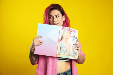 Photo of Beautiful woman with pink dyed hair holding sports magazine on yellow background