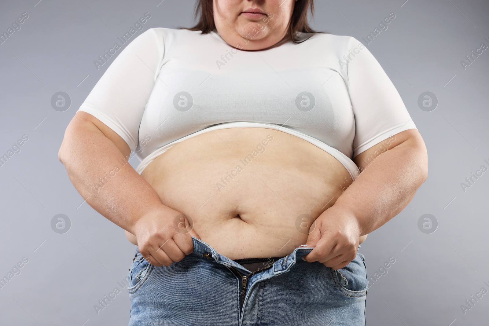 Photo of Overweight woman trying to put on tight jeans against grey background, closeup