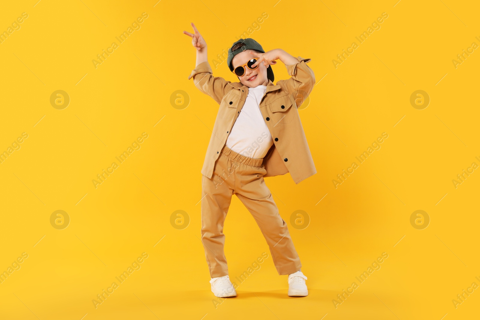 Happy little boy in sunglasses and baseball hat dancing on orange background Photo of Happy little boy in sunglasses and baseball hat dancing on orange background
