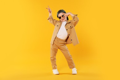 Photo of Happy little boy in sunglasses and baseball hat dancing on orange background