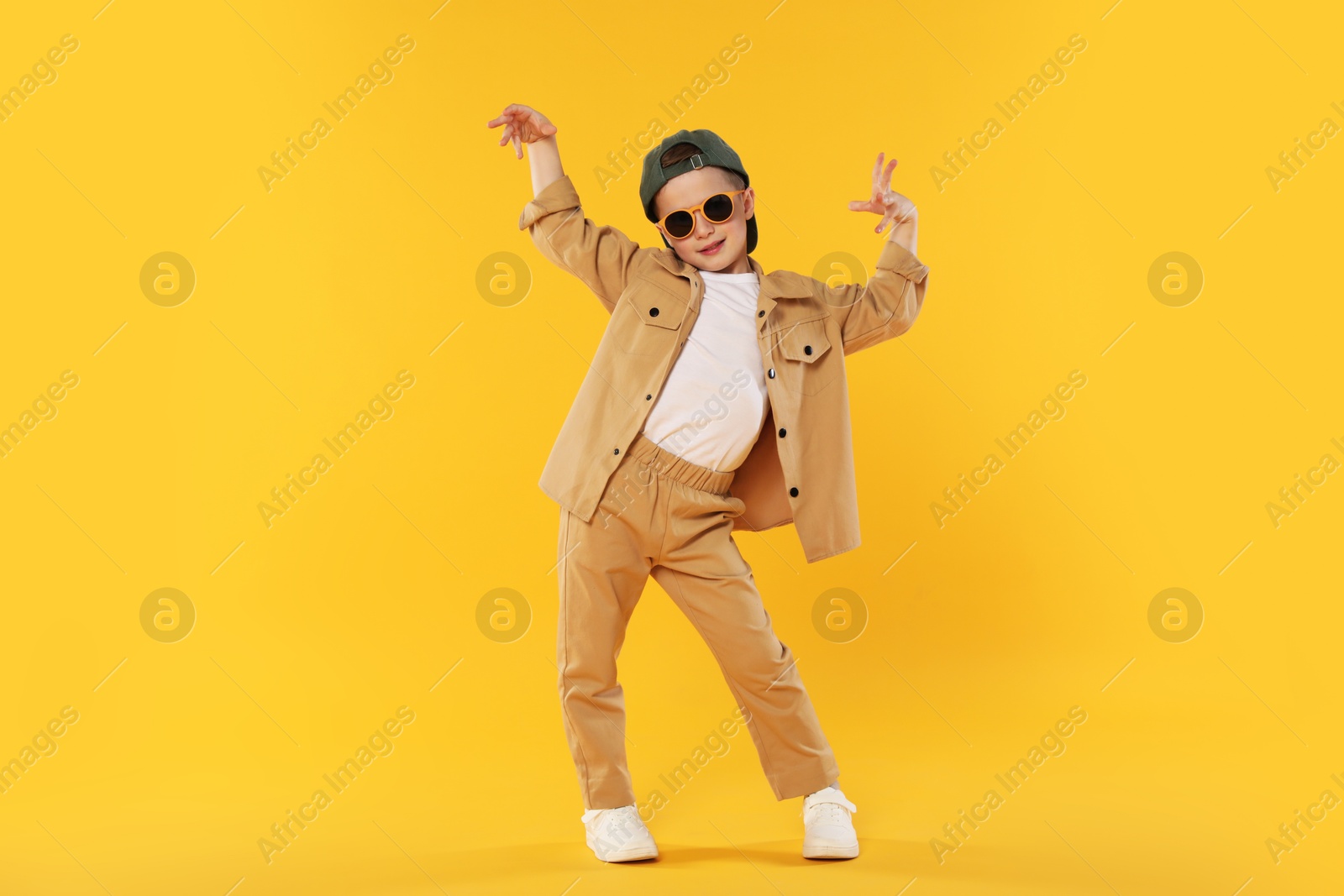 Photo of Cute little boy in sunglasses and baseball hat dancing on orange background