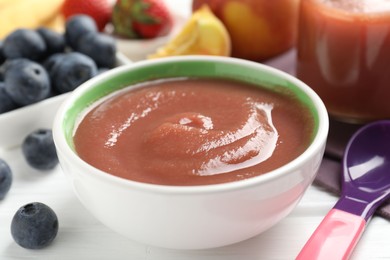 Photo of Baby food. Tasty puree in bowl, spoon and ingredients on white wooden table, closeup