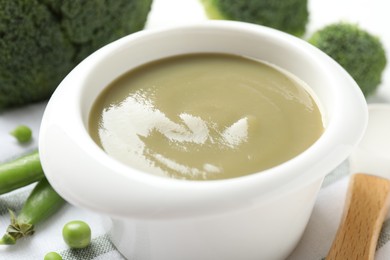 Photo of Baby food. Tasty puree in bowl, spoon, peas and broccoli on white table, closeup