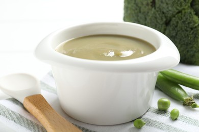 Photo of Baby food. Tasty puree in bowl, spoon, peas and broccoli on white table, closeup