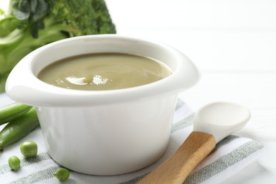 Photo of Baby food. Tasty puree in bowl, spoon, peas and broccoli on white table, closeup
