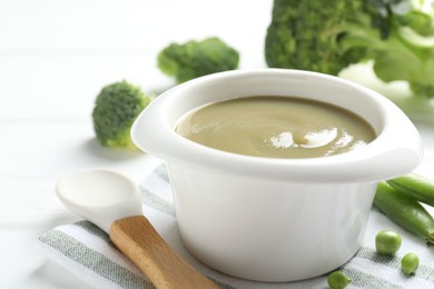 Photo of Baby food. Tasty puree in bowl, spoon, peas and broccoli on white table, closeup