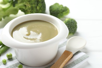 Photo of Baby food. Tasty puree in bowl, spoon, peas and broccoli on white wooden table, closeup