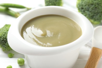 Photo of Baby food. Tasty puree in bowl, spoon, peas and broccoli on white wooden table, closeup