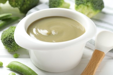 Photo of Baby food. Tasty puree in bowl, spoon, peas and broccoli on white wooden table, closeup