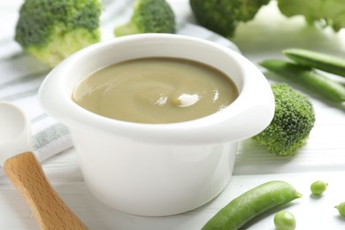 Photo of Baby food. Tasty puree in bowl, spoon, peas and broccoli on white wooden table, closeup