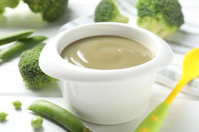 Photo of Baby food. Tasty puree in bowl, spoon, peas and broccoli on white wooden table, closeup