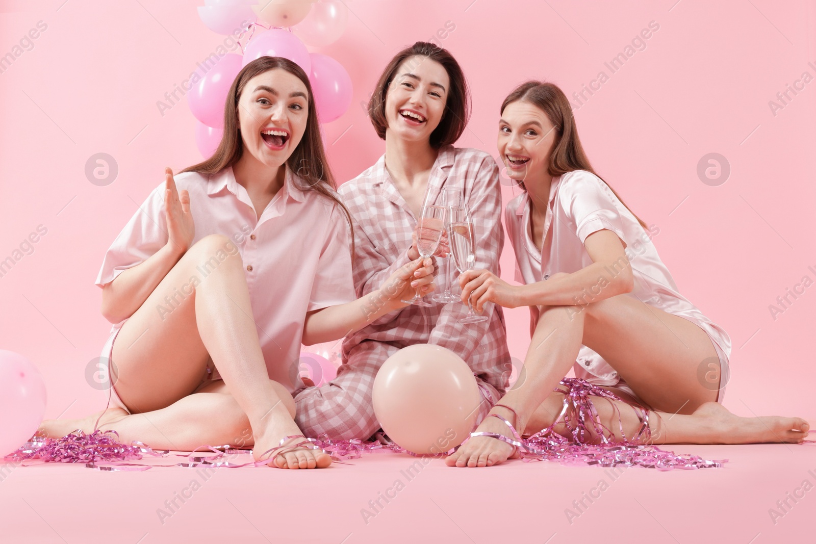 Photo of Pajama party. Happy friends with balloons and tinsel clinking glasses on pink background