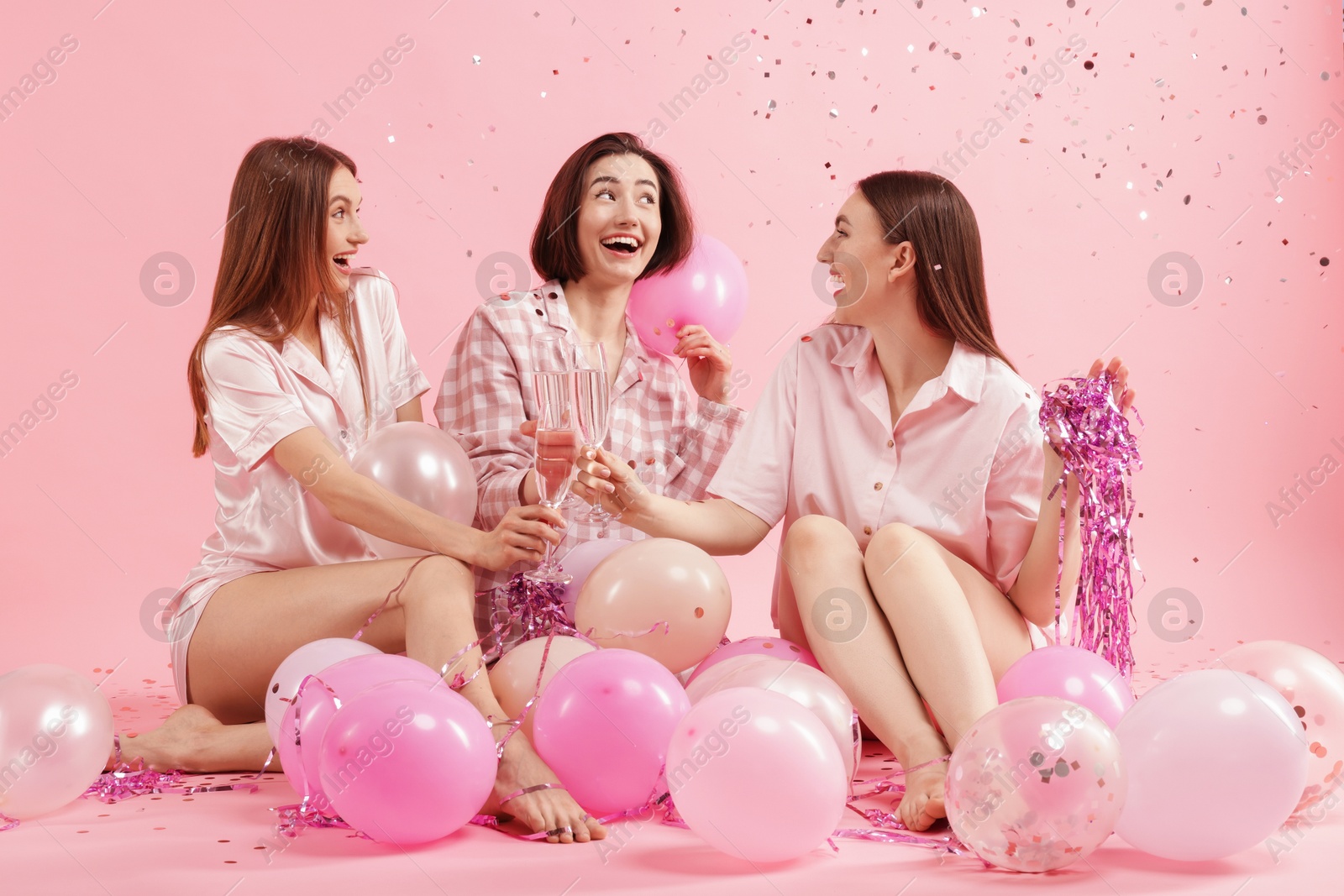 Pajama party. Happy friends with balloons and tinsel clinking glasses under falling confetti on pink background Photo of Pajama party. Happy friends with balloons and tinsel clinking glasses under falling confetti on pink background