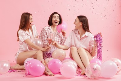 Photo of Pajama party. Happy friends with balloons and tinsel clinking glasses under falling confetti on pink background