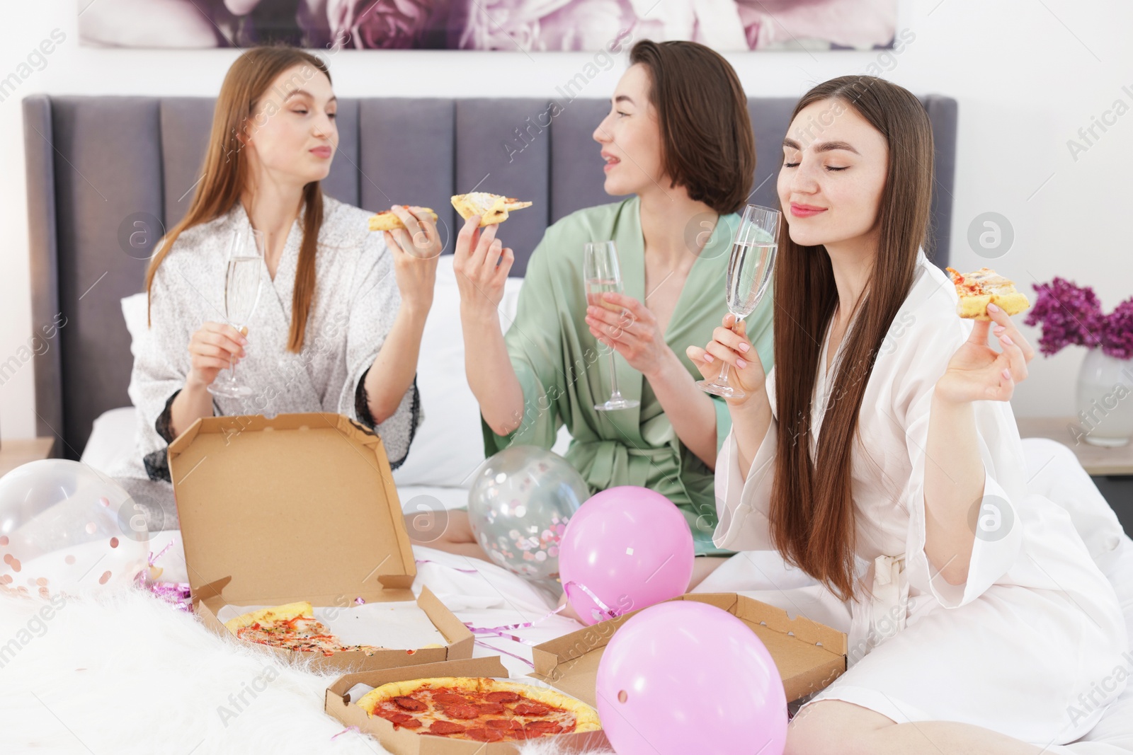 Pajama party. Happy friends with glasses eating pizza on bed at home Photo of Pajama party. Happy friends with glasses eating pizza on bed at home
