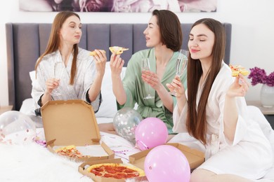 Photo of Pajama party. Happy friends with glasses eating pizza on bed at home