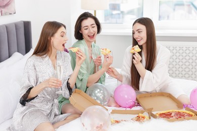 Photo of Pajama party. Happy friends with glasses eating pizza on bed at home
