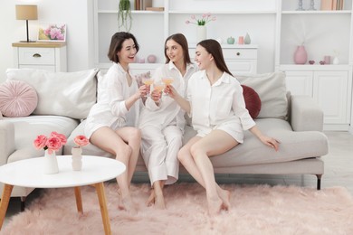 Photo of Pajama party. Happy friends clinking glasses on sofa at home