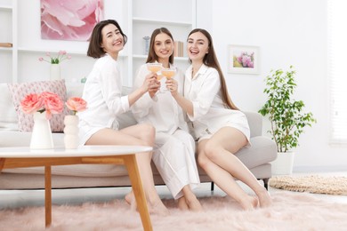 Photo of Pajama party. Happy friends clinking glasses on sofa at home
