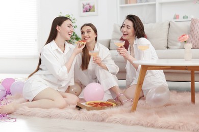 Photo of Happy friends in pajamas with glasses of alcoholic drink eating pizza on floor at home