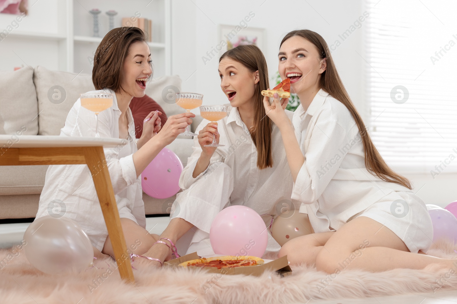 Happy friends in pajamas with glasses of alcoholic drink eating pizza on floor at home Photo of Happy friends in pajamas with glasses of alcoholic drink eating pizza on floor at home