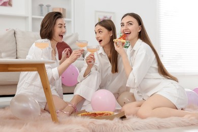 Photo of Happy friends in pajamas with glasses of alcoholic drink eating pizza on floor at home