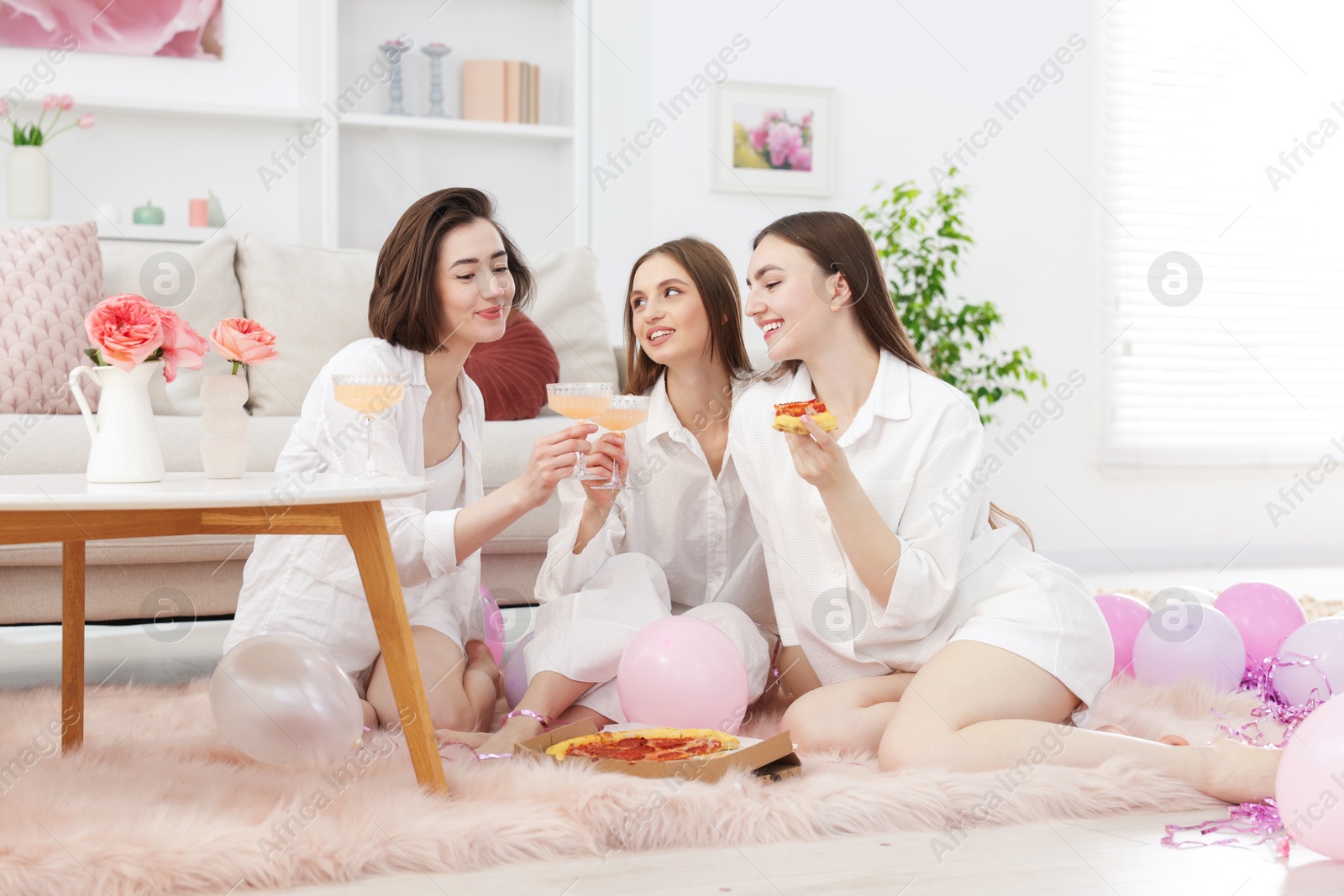 Happy friends in pajamas with glasses of alcoholic drink eating pizza on floor at home Photo of Happy friends in pajamas with glasses of alcoholic drink eating pizza on floor at home