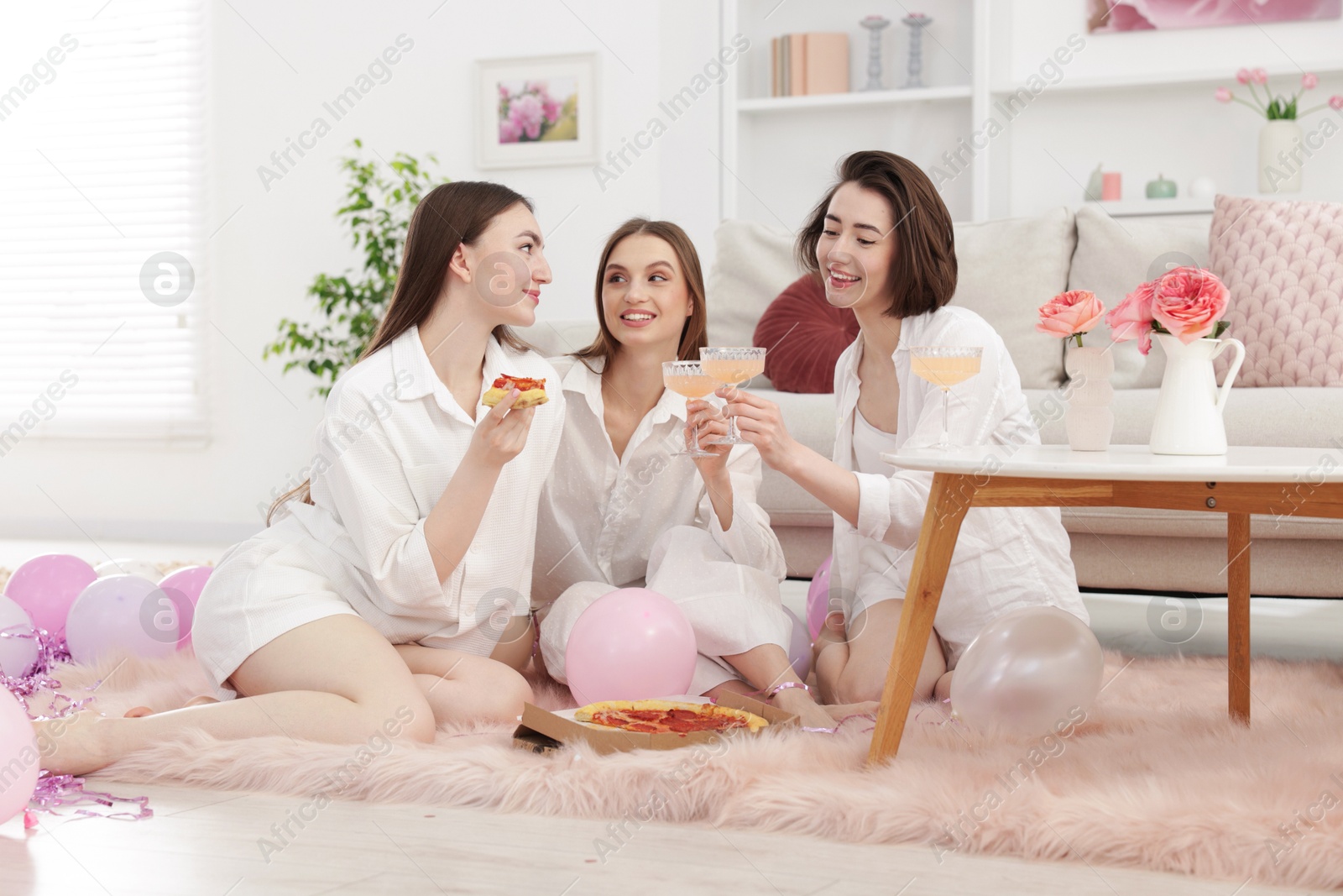 Happy friends in pajamas with glasses of alcoholic drink eating pizza on floor at home Photo of Happy friends in pajamas with glasses of alcoholic drink eating pizza on floor at home