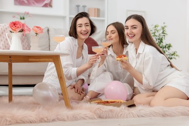 Photo of Happy friends in pajamas with glasses of alcoholic drink eating pizza on floor at home
