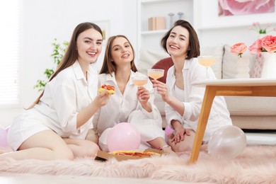 Photo of Happy friends in pajamas with glasses of alcoholic drink eating pizza on floor at home